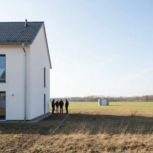 Planer blicken von der scharfen Rückfassade des letzten Hauses über die Abrisskante der Siedlung auf eine weite, leere Wiese.