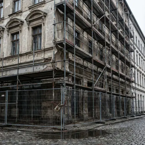 Ein massives Stahlgerüst umschließt vollständig die verlassene, verwitterte Fassade eines Altbaus.