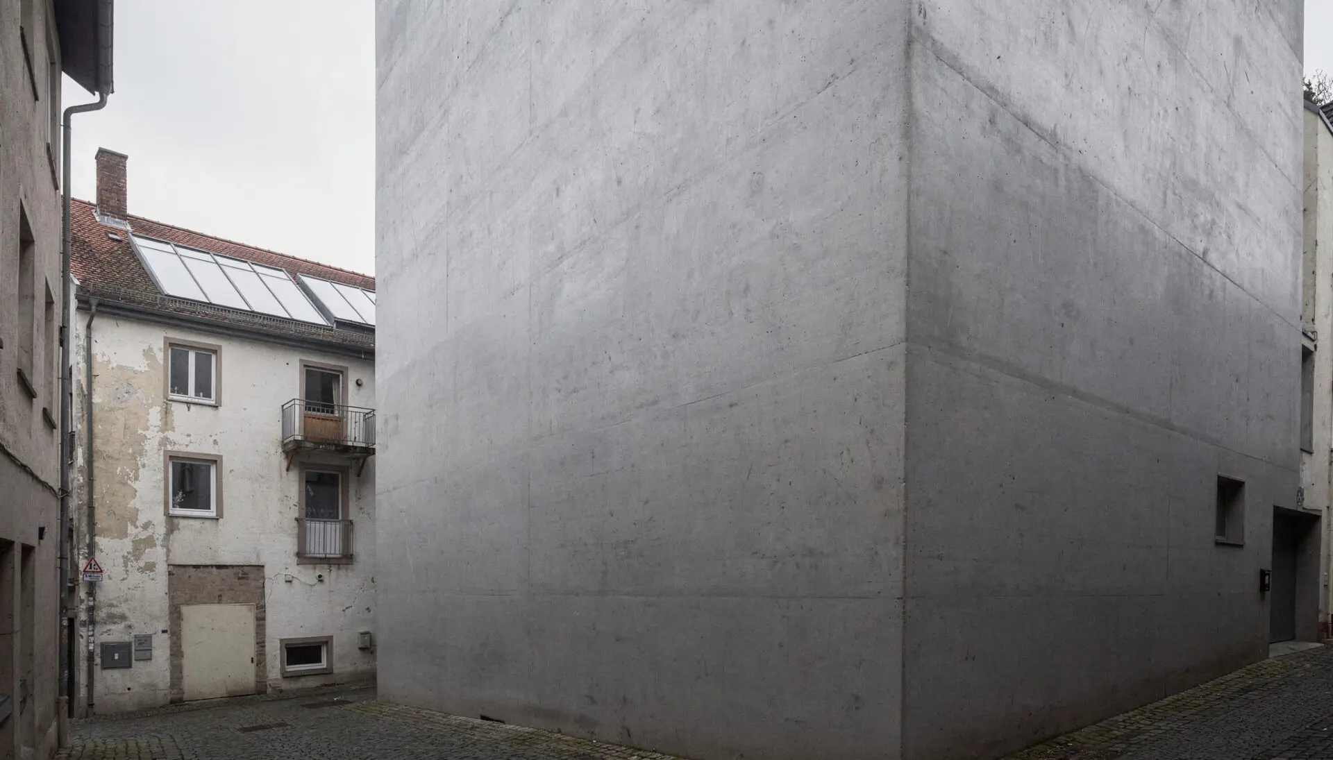 Eine riesige graue Betonwand überragt ein kleines historisches Haus mit Balkon in einer engen Altstadtgasse.